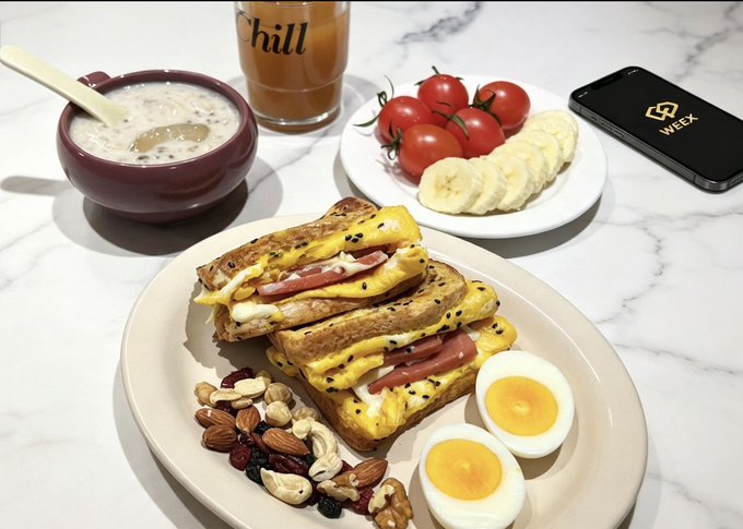 A breakfast spread on a marble countertop featuring egg and ham sandwiches on seeded bread, a hard-boiled egg cut in half, mixed nuts and dried fruit, a bowl of yogurt, sliced bananas, cherry tomatoes, and a glass marked 'chill'.