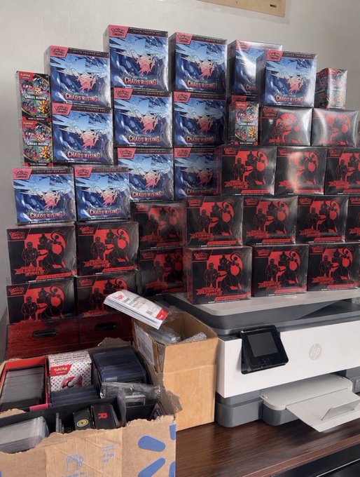 A large collection of Disney Lorcana trading card game 'The First Chapter' booster boxes and booster packs stacked on and around an HP printer.
