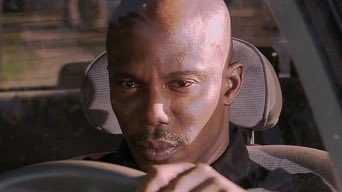 A close-up shot shows a man with a serious expression sitting in the driver's seat of a car.