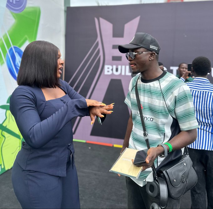 A woman in a navy blue outfit speaks with a young man wearing a green and white striped shirt, sunglasses, and a cap at an event with "BUILD" visible on the background signage.