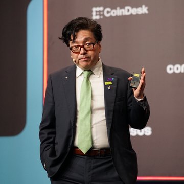 A man in a navy suit with a green tie stands on stage holding up a smartphone, with the "CoinDesk" logo visible in the background. The image relates to the post as it appears to feature Tom Lee, the analyst cited as commenting on Ethereum's strong Q3 performance and its influence on crypto-related stocks in the Russell 1000.