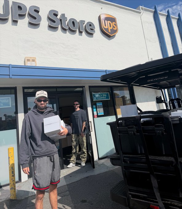 Two men standing outside a UPS Store location; one is holding a white box, with a black utility vehicle parked on the right.