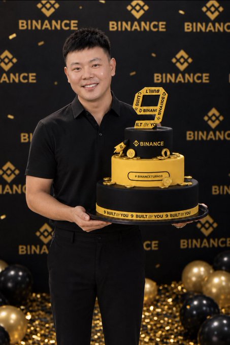 A man in a black shirt holding a tiered yellow and black Binance-branded 9th anniversary cake, with Binance logos in the background.