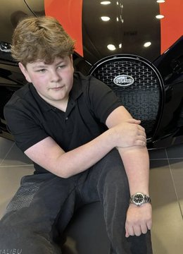A young teen boy sits casually in front of a black Bugatti with the brand's logo visible on the front grille, wearing a black polo shirt and a silver wristwatch. The caption connects to the post's claims about the "Quant Kid," depicting the 15-year-old alleged crypto/memecoin trader in a setting associated with the lavish lifestyle the post describes.