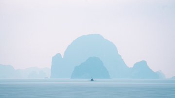 A small boat floats on calm misty water with large karst rock formations rising in the hazy background.