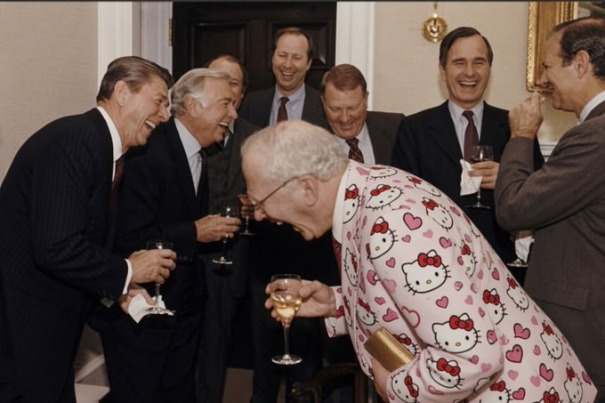 A photograph of several formally dressed men laughing, with one older man in the center wearing a pink Hello Kitty patterned suit and holding a wine glass.