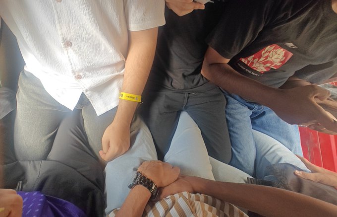 An overhead shot of several people sitting closely together, showing their laps, hands, and clothing, including someone wearing a yellow wristband and another with a watch.
