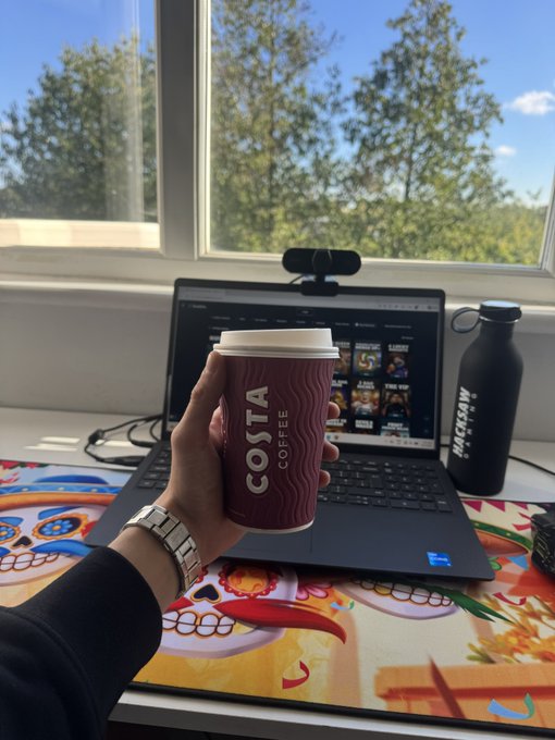 A person's hand holds a Costa Coffee cup while seated at a desk with an open laptop, next to a black water bottle, with trees visible through the window behind.