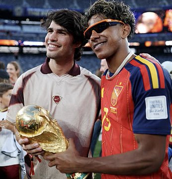 Two smiling soccer players pose together, with one holding the FIFA World Cup trophy; one wears a maroon and white kit and the other wears a red Spain national team jersey.