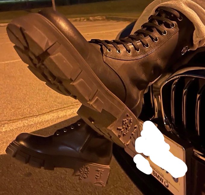 A person rests their rugged black combat boots on the rear of a BMW at night, with the license plate partially obscured, playfully tying the pricey luxury car to the question "How much $SOL?"—suggesting the cost could buy a whole vehicle.