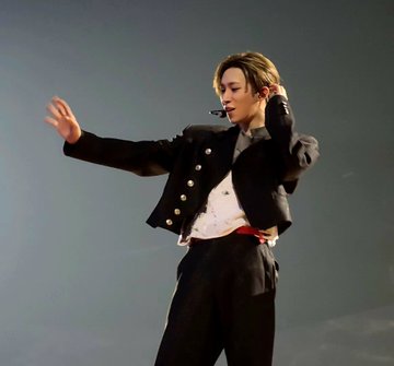 A performer in a black military-style jacket and white shirt dances on stage while wearing a headset microphone against a gray backdrop.