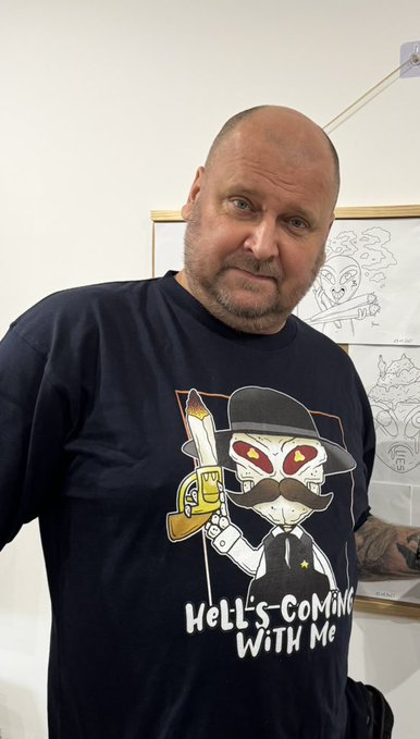 A bald man with a short beard poses wearing a navy t-shirt featuring a cartoon luchador graphic and the text 'HeLL'S-COMiNG WiTH Me'.