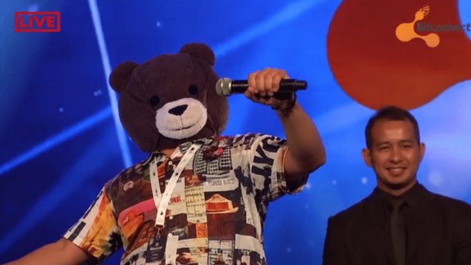 A live broadcast screenshot showing a person wearing a brown teddy bear mask holding a microphone on stage, with another person in a black suit smiling behind them and a 'LIVE' indicator in the corner.
