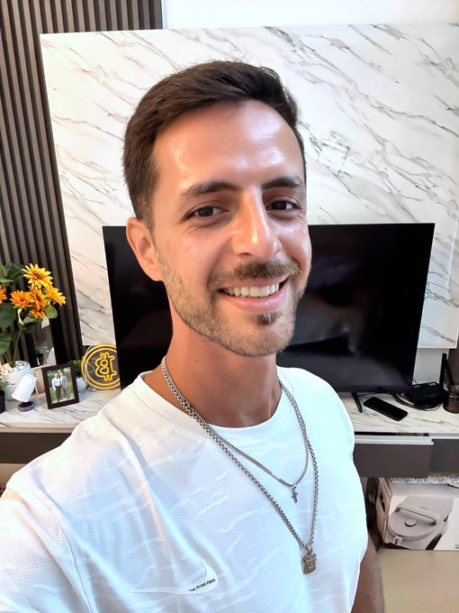 A smiling man with short brown hair and a beard takes a selfie indoors, wearing a white textured t-shirt and layered silver necklaces, with a marble-patterned wall and TV in the background.