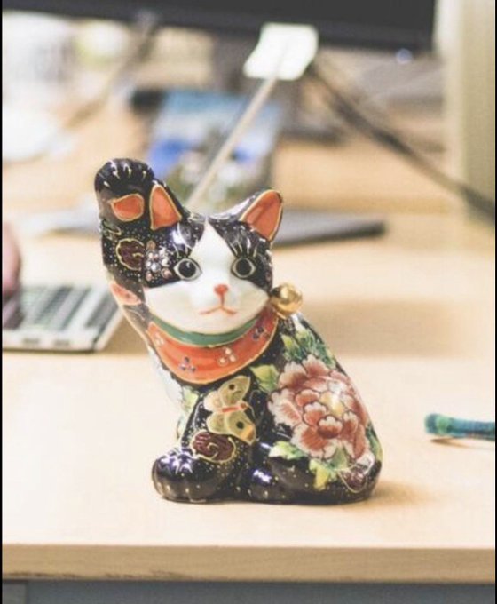 A black ceramic Maneki-neko (Japanese lucky cat) figurine decorated with floral patterns and a red collar with a bell, sitting on a desk with a laptop and pencil in the background.