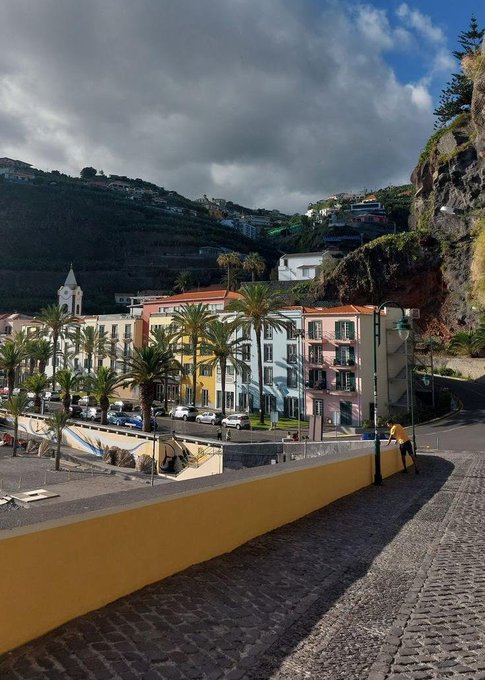 A coastal town square with pastel-colored buildings, palm trees, and a church nestled against a steep green hillside, viewed from a cobblestone path under dramatic cloudy skies—setting a serene, early-morning scene that perfectly complements the "GM" (good morning) greeting.