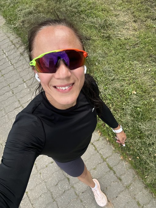 A woman smiles at the camera while taking a selfie during a run, wearing reflective sunglasses, a black athletic top, and a white smartwatch on a paved path beside a grassy area—capturing the active lifestyle that pairs well with the post's mention of a running day and STEPN tickets.