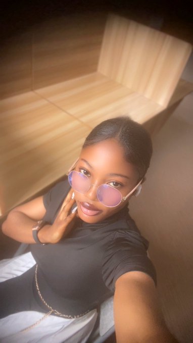 A selfie of a woman wearing round purple-tinted sunglasses and a black top, posing in what appears to be a restaurant or lounge with wooden paneling in the background.