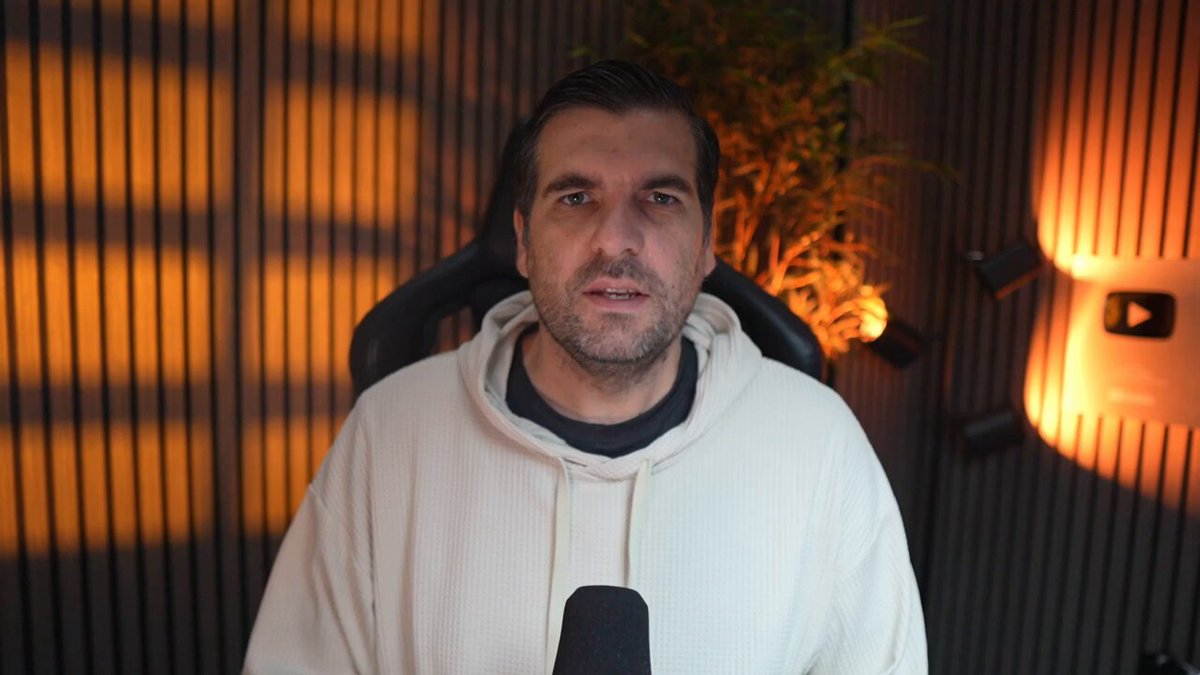 The image depicts a man wearing a white hoodie, seated in front of a large window with sunlight streaming through. The man appears to be engaged in an interview or discussion, possibly in a conference room setting. He is speaking into a microphone attached to a boom mic stand. His facial expression suggests he may be serious about the topic being discussed.

The background includes a plant and some lights that could possibly indicate a special event or formal setting for the gathering. The image also contains text related to Wall Street, which might be connected to the man's story or context.