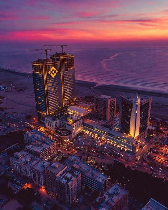 An aerial sunset view of a coastal city featuring a prominent building displaying the Binance (BNB) logo, with construction cranes visible nearby.