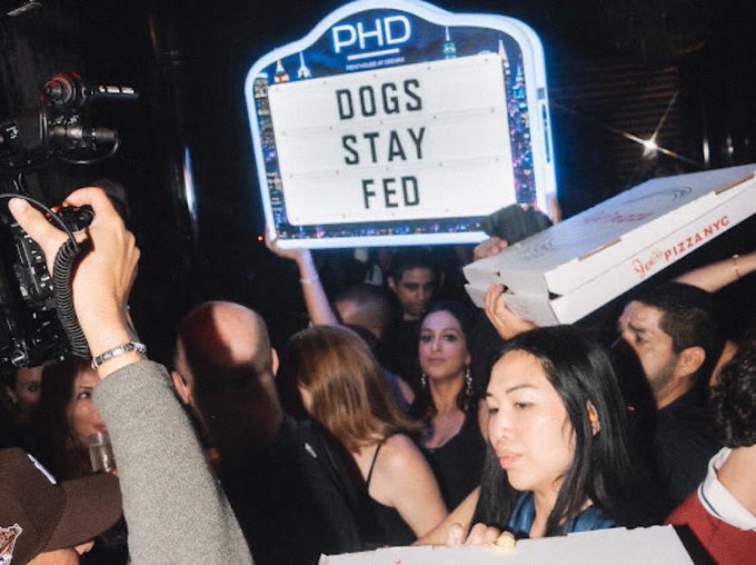 A crowded nightclub or party scene where someone is holding up a neon-style sign reading "PHD DOGS STAY FED", while a person in the foreground holds a "Soho Pizza NYC" pizza box and another films with a camera.