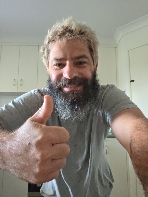 A selfie of a smiling, bearded man with messy blonde hair giving a thumbs-up, wearing a gray t-shirt in what appears to be a kitchen.