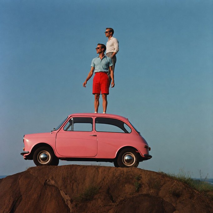 Two men wearing sunglasses, polo shirts, and shorts stand stacked on top of a small pink Fiat car perched on a dirt mound against a clear blue sky.