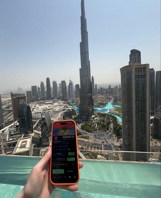 A phone displaying crypto trading apps with "$38,928.38" and "Referee" text is held up against a luxurious Dubai skyline featuring the Burj Khalifa, capturing the aspirational "Memecoin trader lifestyle" promised by crypto profits. The juxtaposition of the modest trading interface with the high-rise cityscape symbolizes the wealth dreams fueling memecoin speculation.