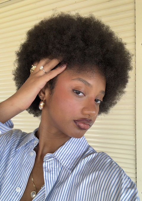 A selfie of a woman with a natural afro hairstyle wearing a blue and white striped shirt, gold rings, and earrings, posing with her hand near her face.