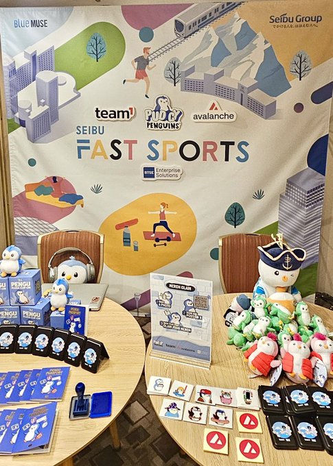 A promotional table display for 'Pudgy Penguins' at a 'Seibu Fast Sports' event, featuring branded merchandise like plush toys, keychains, and stickers arranged on a table with an illustrated backdrop.