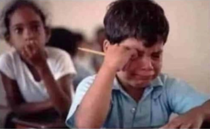 A blurry, low-quality image of a young boy in a blue shirt crying and rubbing his eye, with a young girl visible in the background.