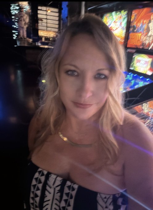 A selfie of a blonde woman wearing a black and white patterned top and gold necklace in a dimly lit arcade with pinball machines and neon signs visible behind her.