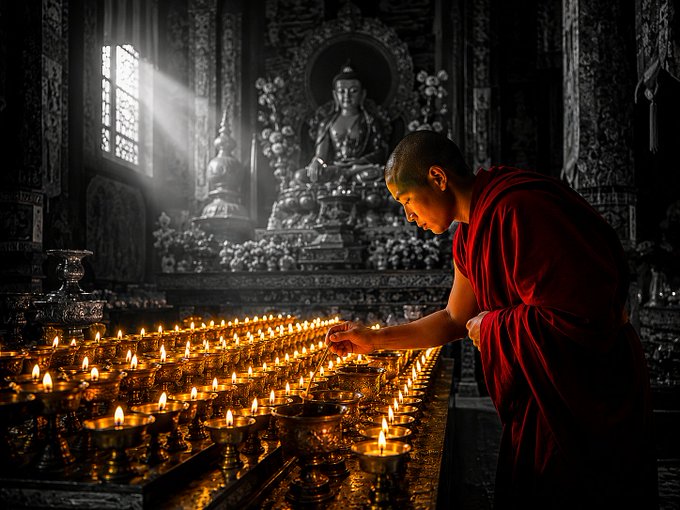 A young Buddhist monk in a crimson robe lights rows of butter candles inside a dimly-lit temple, with a large Buddha statue illuminated in the background.