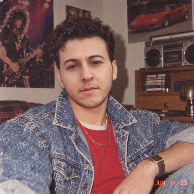 A vintage photo dated 'JUN 14 85' shows a young man with curly dark hair wearing a denim jacket over a red shirt, posing in a room with rock posters and a boombox.