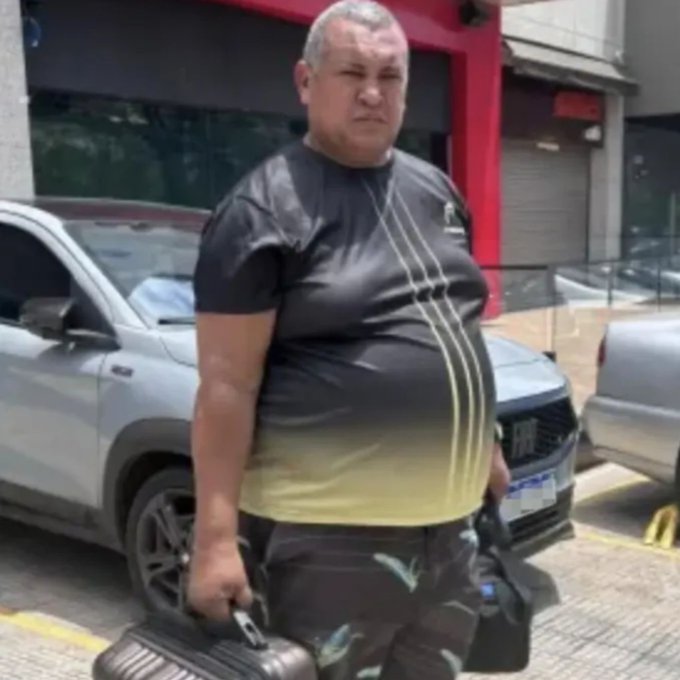 A muscular man standing outdoors near parked cars, carrying a rolling suitcase and wearing a black athletic shirt and patterned shorts.
