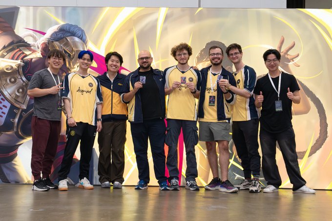 A group photo of seven men, several wearing matching yellow and navy blue esports jerseys with 'Booyah!' text, posing in front of a game-themed promotional backdrop.