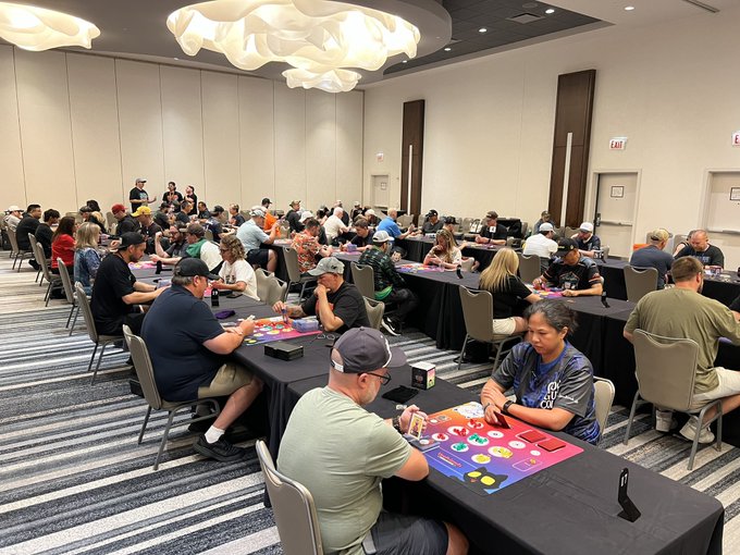 Large gathering in a ballroom where many people are seated at long tables playing board games and card games.