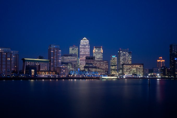 A nighttime cityscape of London's Canary Wharf skyline, with its illuminated skyscrapers reflecting across the River Thames. The gleaming financial district visually evokes the concept of "Tokenization," representing the digitization and transformation of traditional assets in the modern financial world.