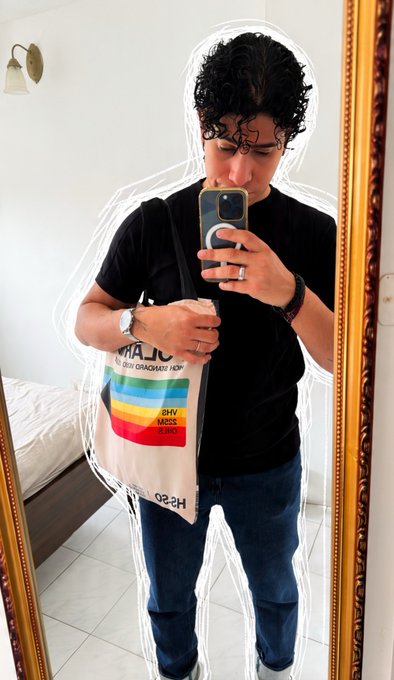 A mirror selfie of a man wearing a black t-shirt and blue jeans, holding a phone, with a tote bag slung over his shoulder that displays a rainbow graphic with the text 'HIV AIDS' and 'HS 20'.