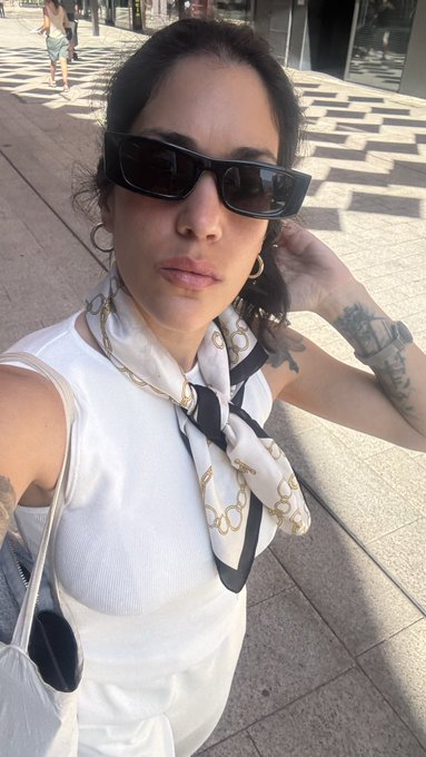 A woman takes a selfie outdoors on a tiled pavement, wearing a white sleeveless top, dark sunglasses, a chain-patterned silk scarf, and hoop earrings.