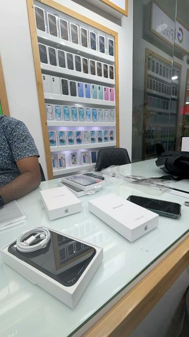 A customer sits at a glass display counter in what appears to be a phone shop called "SKYNET," with open iPhone boxes and accessories on the counter, surrounded by walls displaying rows of smartphones—tying in with the post about making a small purchase before getting back to work ("lock in").