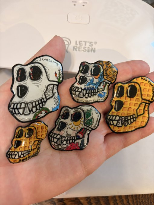 A hand displays five colorful sugar skull-shaped pins or brooches featuring intricate designs, held over a white device labeled 'LET'S RESIN'.