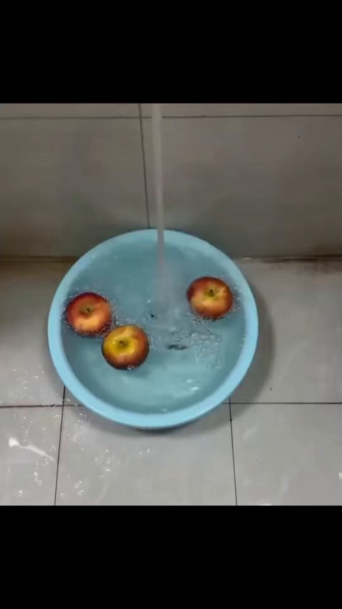 The image features a blue bowl filled with water, containing three apples floating inside of it. The bowl is placed on a tiled floor, and there are two other bowls visible in the scene - one located near the top left corner and another at the bottom right corner.