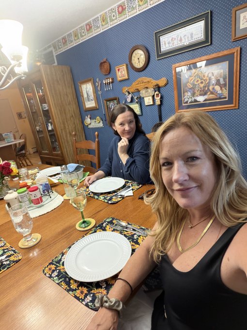 A selfie-style photo of two women seated at a wooden dining table set with plates and floral placemats in a warmly decorated room with blue walls and framed artwork.