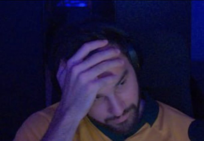 A man with a beard wearing headphones and a yellow shirt holds his hand to his forehead in a dimly lit blue room, appearing frustrated or tired.