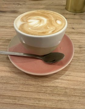 A cappuccino with latte art served in a white cup on a pink saucer with a spoon on a wooden table.