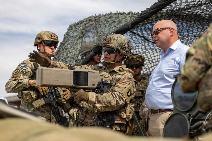 Military personnel in camouflage uniforms and helmets gather around tactical equipment under camouflage netting, while a bald man in sunglasses and a light blue button-down shirt stands nearby observing.