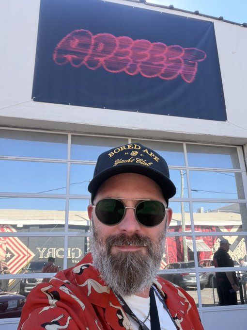 A man wearing a "Bored Ape Yacht Club" cap and sunglasses poses in front of a large BAYC logo banner at ApeFest Charleston, embodying the event's signature style with his red shirt and rugged look.
