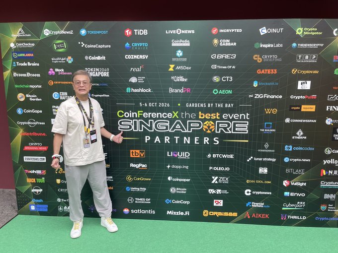A man wearing glasses and a white shirt poses at the 'CoinFerenceX SINGAPORE PARTNERS' event, held on 5-6 OCT 2024 at Gardens by the Bay, standing in front of a backdrop covered in numerous cryptocurrency company logos.