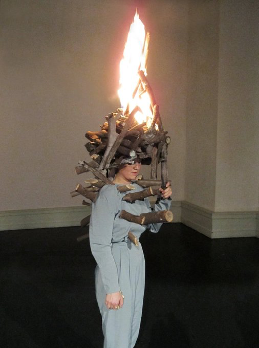 A woman in a light blue outfit stands in a gallery space, wearing a sculptural headdress made of stacked wooden logs with a large flame burning from the top.